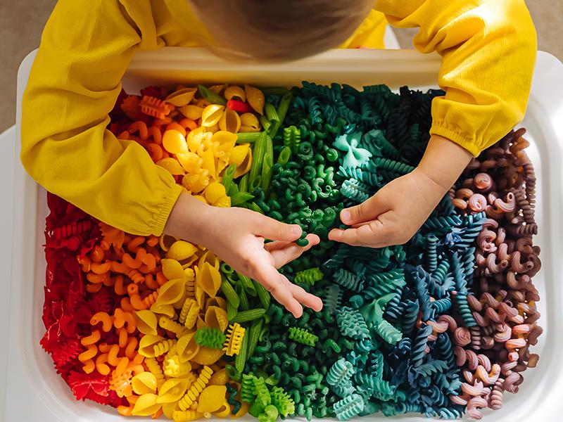 Child leaning over bin of multicolored pasta